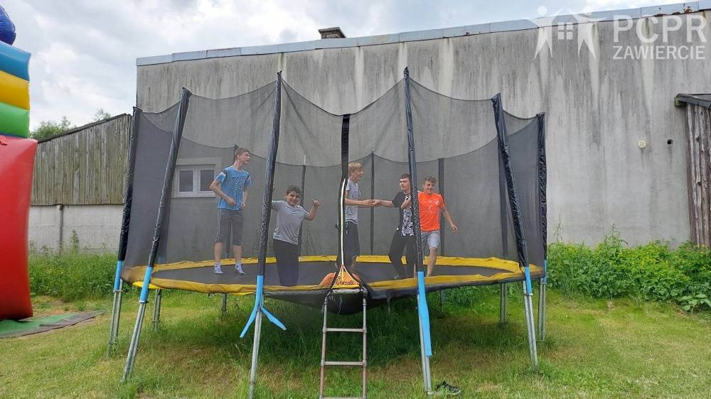 Zdjęcie: Pięciu chłopców skacze na dużej trampolinie na trawie, są rozbawieni. W tle widać budynek, po lewej stronie kawałek dmuchanego zamku.